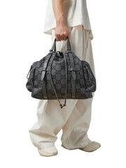 A person wearing cream-colored draped clothing holds a dark gray patterned shoulder bag with double top handles and a long strap, photographed against a clean white studio background.