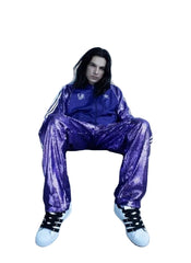 Person wearing a purple Adidas tracksuit sitting on a tire against a white wall.