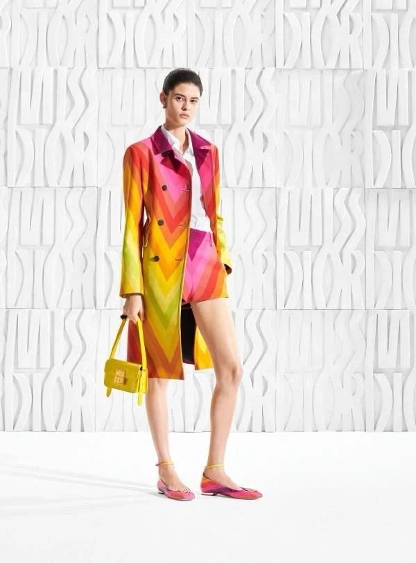 A model stands against a white textured wall wearing a vibrant, multicolored geometric print trench coat and matching shorts, holding a small yellow handbag.
