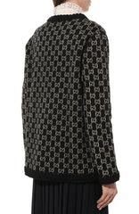 A person shown from the back wearing a black knit sweater with an allover beige interlocking monogram pattern and a black lace-trim collar, paired with a pleated black skirt against a plain white background.