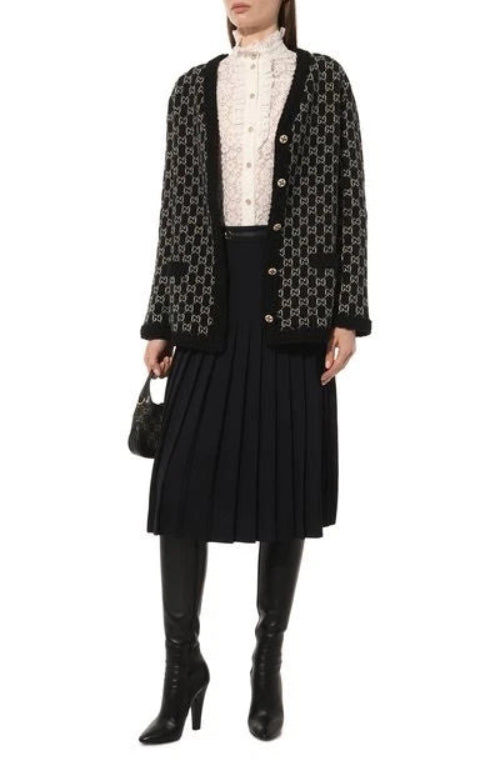 A model wears a black and white GG monogram tweed cardigan over a white lace mock-neck top, paired with a black pleated skirt, black knee-high leather boots, and a small black handbag against a plain white background.
