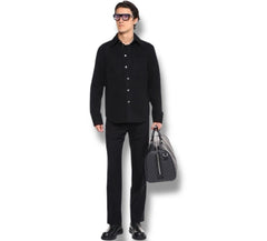 A man wearing a black long-sleeve button-up shirt, black trousers, and black shoes, holding a black patterned duffel bag against a plain white background.
