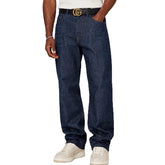 A person wearing dark blue denim jeans with a gold-tone belt and white sneakers, shown from the waist down against a plain light gray studio background.