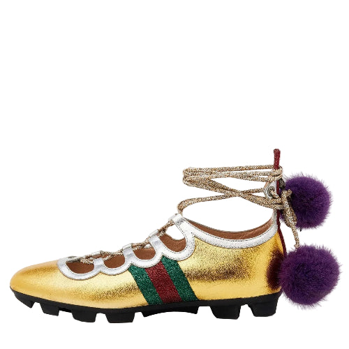 A metallic gold leather shoe with a green and red web stripe, scalloped white leather trim, lace-up ankle detailing, and two purple faux fur pom-poms hanging from the heel, shown against a plain light gray background.