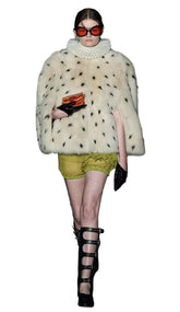 Model wearing a white fur coat with black spots, green shorts, and black high-heeled shoes on a runway.
