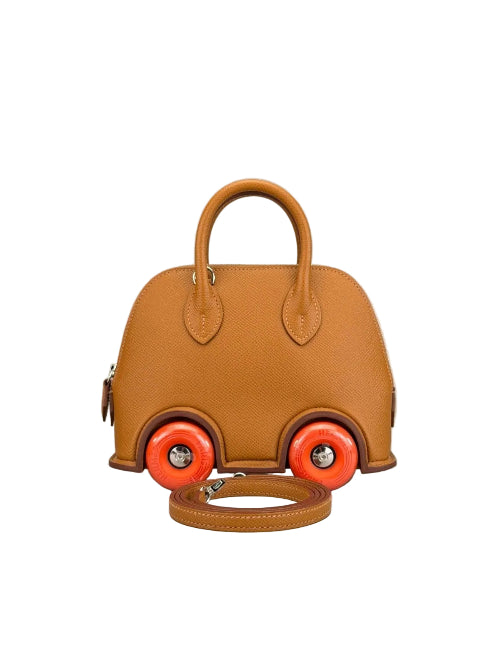 A structured tan leather handbag designed to look like a small car, featuring bright orange wheels with silver hubcaps and a matching tan leather shoulder strap resting in front, set against a neutral gray background.