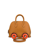 A structured tan leather handbag designed to look like a small car, featuring bright orange wheels with silver hubcaps and a matching tan leather shoulder strap resting in front, set against a neutral gray background.