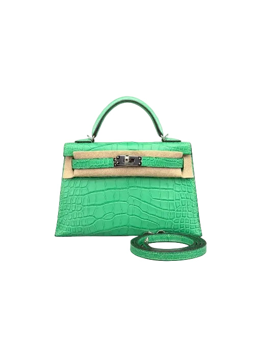 A small structured handbag in bright green crocodile-embossed leather with a top handle, front flap, silver-tone hardware, and a matching green shoulder strap placed in front, photographed on a light gray studio background.