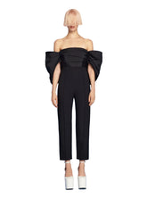 Black off-shoulder jumpsuit with a large bow detail on a blue background