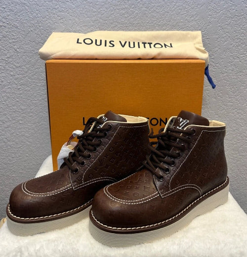A pair of Louis Vuitton brown embossed leather ankle boots with white soles, displayed in front of their signature orange Louis Vuitton box and beige dust bag.