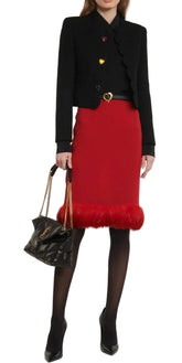 A model wearing a black cropped jacket with gold heart buttons, a red pencil skirt with a red faux-fur hem, black tights, and holding a black quilted handbag with a gold chain strap.