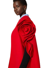 A model shown from the side wearing a bright red cape-style coat with a large sculptural rose detail on the shoulder, layered over a black sleeve and a light blue collared shirt, against a plain light beige background.