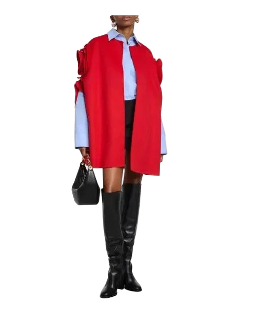 A model wearing a bright red oversized wrap-style coat with dramatic shoulder ruffles, layered over a light blue collared shirt, black skirt, black knee-high boots, and holding a black handbag against a light gray studio background.