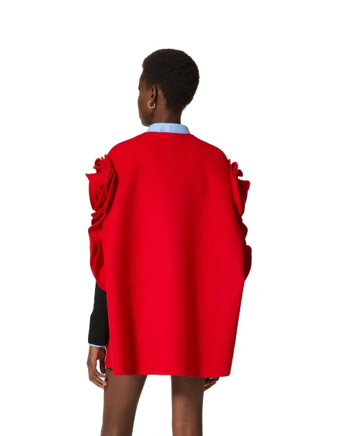 A model shown from the back wearing a bright red cape-style jacket with dramatic sculptural ruffle details on the shoulders, layered over a light blue collared shirt and a black long-sleeve top, against a solid off-white background.