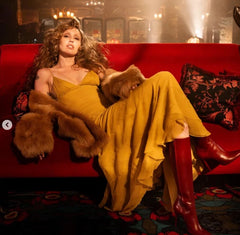 A woman reclines on a red sofa wearing a mustard yellow dress, brown fur trim, and red knee-high boots in a dimly lit, atmospheric interior.