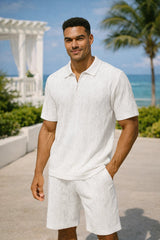 Man wearing a white polo shirt and shorts standing on a path with ocean and palm trees in the background.
