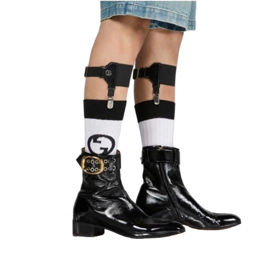 Person wearing black leather boots with buckles, white socks, and black suspenders on a neutral background.