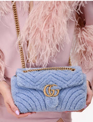 Blue textured handbag with gold chain and logo held by a person wearing a pink feathered garment.