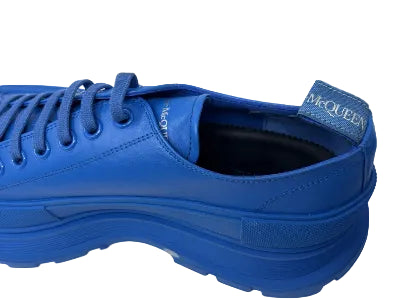 A close-up side view of a McQueen low-top sneaker in a vibrant royal blue leather, featuring matching blue laces, a thick textured sole, and a blue heel pull tab with the McQueen logo.