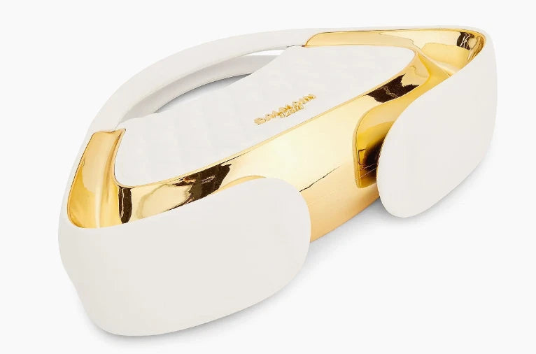 A white and gold metallic visor-style headband or mask with a glossy reflective finish, shown on a plain light gray background. The brand name 'BLOOMBERG' is printed in gold on the white inner surface.