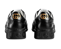 A pair of black leather low-top sneakers shown from the back, featuring a prominent interlocking GG logo in gold-tone metal on the heel counters.
