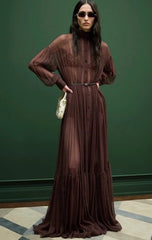 A model wearing a long-sleeved, high-neck, button-front maxi dress in a deep brown or burgundy chiffon-like fabric, styled with a matching belt and a small white handbag, set against a dark green wall.