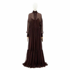 A dark brown long-sleeved maxi dress with a sheer mesh high-neck overlay and a pleated skirt, displayed on a white mannequin against a plain white background.