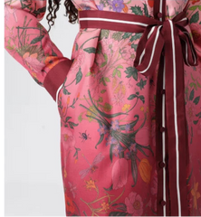 A close-up of a pink floral print dress featuring a dark red waist tie with white piping, a button-down front, and a long sleeve with matching cuffs.