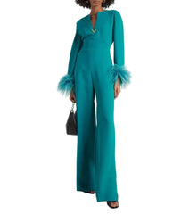 A model wearing a vibrant teal green wide-leg jumpsuit with long sleeves and ruffled cuffs, styled with a black hat and a small black handbag against a plain white background.