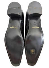 A pair of black leather dress shoes shown from the sole side, positioned in front of their black Saint Laurent box on a beige carpet. The soles feature gold lettering reading 'SAINT LAURENT PARIS' and a gold square logo patch.