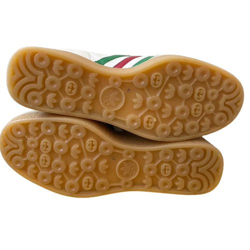 The soles of a pair of white leather sneakers with green and red stripes, showing a tan gum rubber bottom with circular tread patterns.