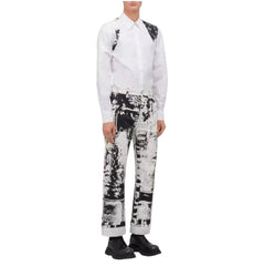 A model wearing a white long-sleeve shirt with black shoulder patches and black-and-white patterned trousers, standing against a plain white background.