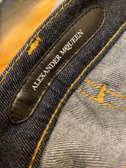 Close-up of a pair of dark blue denim jeans showing the inner waistband label which reads 'ALEXANDER MCQUEEN' in white text, with yellow contrast stitching.