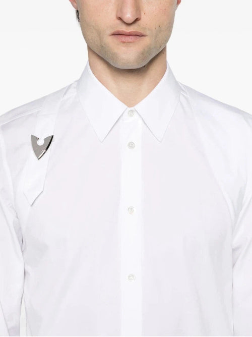 A close-up of a man wearing a white long-sleeve button-down shirt with a unique metallic silver wing-shaped lapel pin on the left chest, set against a plain white background.