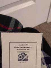 A close-up of a white Legitapp Certificate of Authenticity card held in front of a plaid garment, with a white wall and carpet visible in the background.