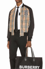 A man wearing a Burberry check jacket over a black shirt and trousers, holding a black Burberry shopping bag with the brand name in white text.