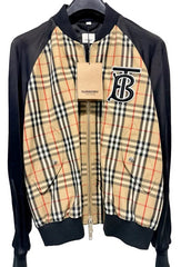 A Burberry zip-up bomber jacket with black ribbed sleeves, collar, and hem, featuring a beige plaid body with red and blue check lines and a large embroidered Burberry logo on the chest.