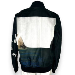 A black bomber jacket shown from the back, featuring a large white rectangular panel on the upper back, a blue and gray landscape graphic across the lower back, and white text on the left sleeve.