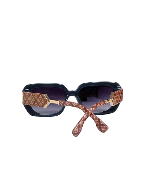 A pair of oversized square sunglasses with dark lenses and a pink patterned nose bridge, resting on a white cylindrical pedestal with a blue patterned box in the background.