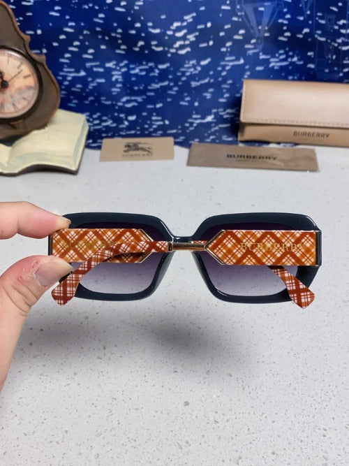 A hand holds a pair of Burberry sunglasses with a black frame and orange-and-white plaid patterned temples, with the Burberry box and case blurred in the background.