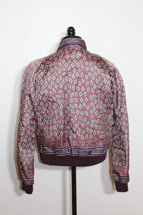 A long-sleeve patterned jacket displayed on a black dress form mannequin against a plain white wall. The jacket features a geometric diamond pattern in dusty pink, white, and dark purple, with a solid dark purple ribbed hem and cuffs.
