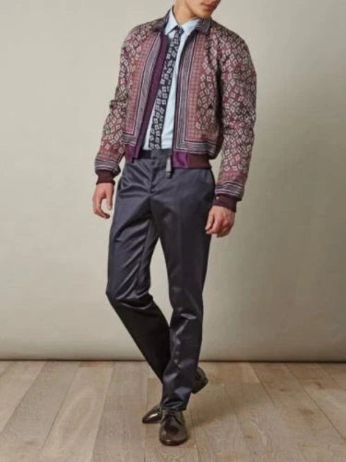 A man wearing a patterned bomber jacket, a blue and white paisley tie, a light blue shirt, and dark charcoal gray trousers, standing against a neutral beige wall and light wood floor.