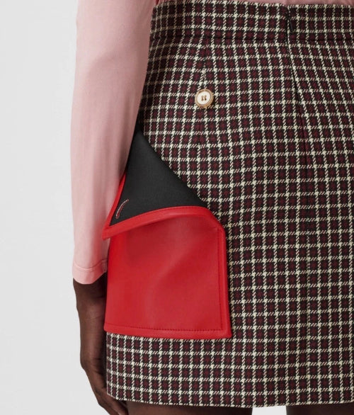 Close-up of a person wearing a brown and cream houndstooth skirt with a large red leather side pocket and a black lining, paired with a light pink long-sleeve top.
