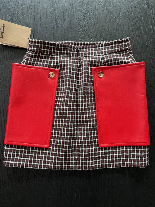 A Burberry mini skirt featuring a brown and white check pattern with two large, vibrant red front flap pockets, displayed flat on a dark wood background with a brand tag visible.