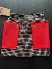 A Burberry mini skirt featuring a brown and white check pattern with two large, vibrant red front flap pockets, displayed flat on a dark wood background with a brand tag visible.
