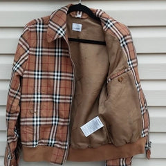 A vintage Burberry plaid jacket in brown, beige, and white, shown open on a black hanger against a white siding background. The jacket features a classic check pattern on the front and sleeves, a solid brown collar, and a brown lining.