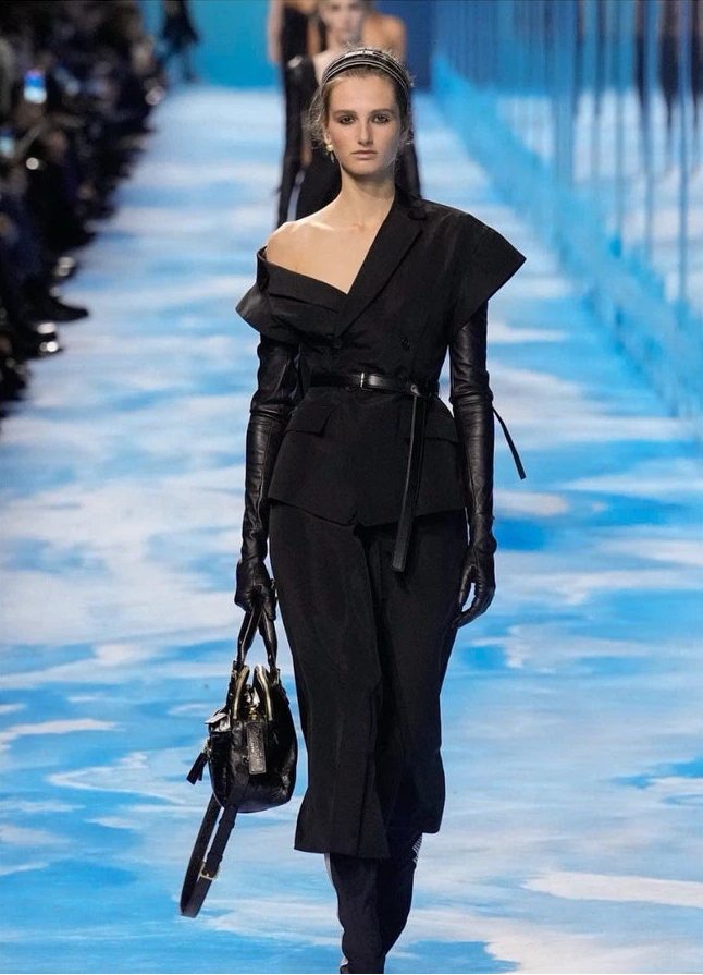 A model walks a fashion runway wearing a black asymmetrical one-shoulder outfit with long black sleeves, a matching belt, and carrying a black handbag against a bright blue runway background.