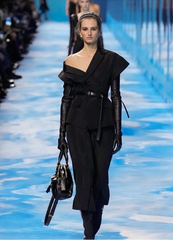 A model walks a fashion runway wearing a black asymmetrical one-shoulder outfit with long black sleeves, a matching belt, and carrying a black handbag against a bright blue runway background.
