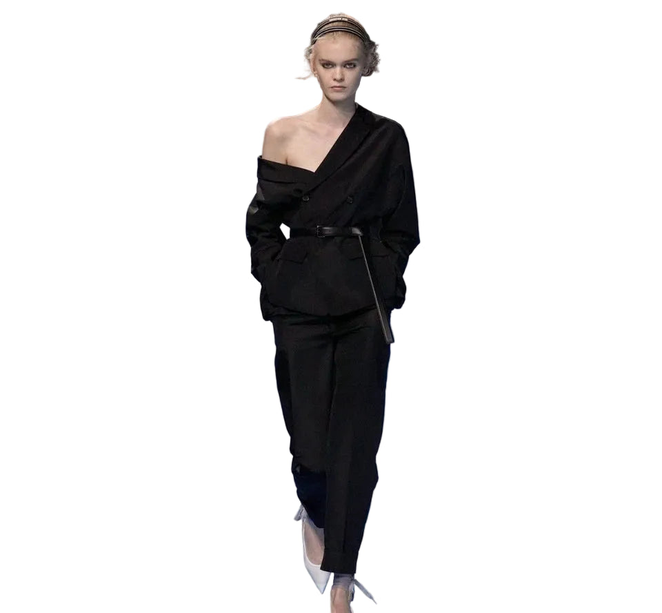 A model walks down a runway wearing a black off-the-shoulder belted one-piece suit with matching trousers, set against a blue cloud-patterned catwalk.