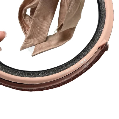 Close-up of a circular cosmetic container with a glossy dark brown translucent lid, a pale pink rim, black textured seal, and a beige satin ribbon tied inside.